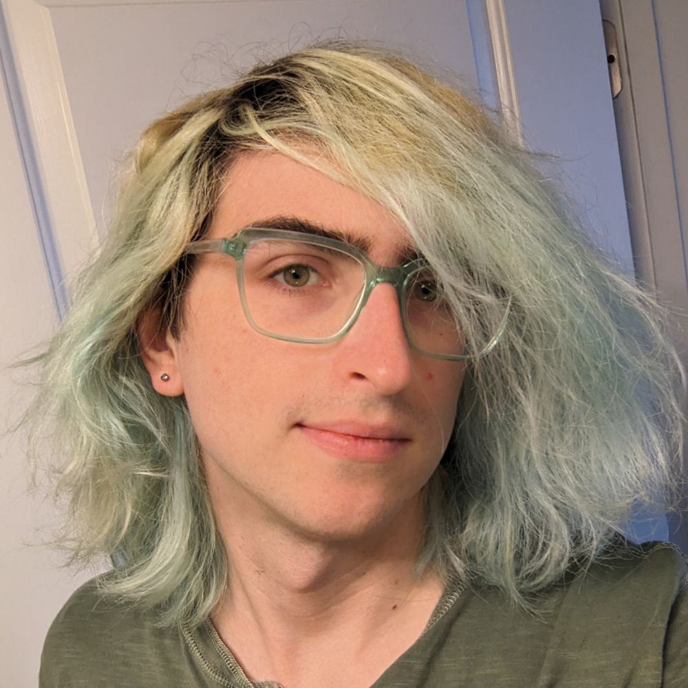selfie of a person with shoulder length faded blue hair, green eyes, blue-green glasses, blue stud earrings, wearing a dark green shirt.
